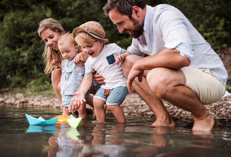 A young family with two toddler children outdoors by the river in summer, playing with paper boats.